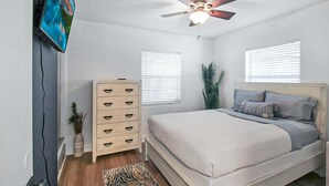 Un dormitorio con una cama, ventilador de techo, cómoda y dos ventanas con persianas.