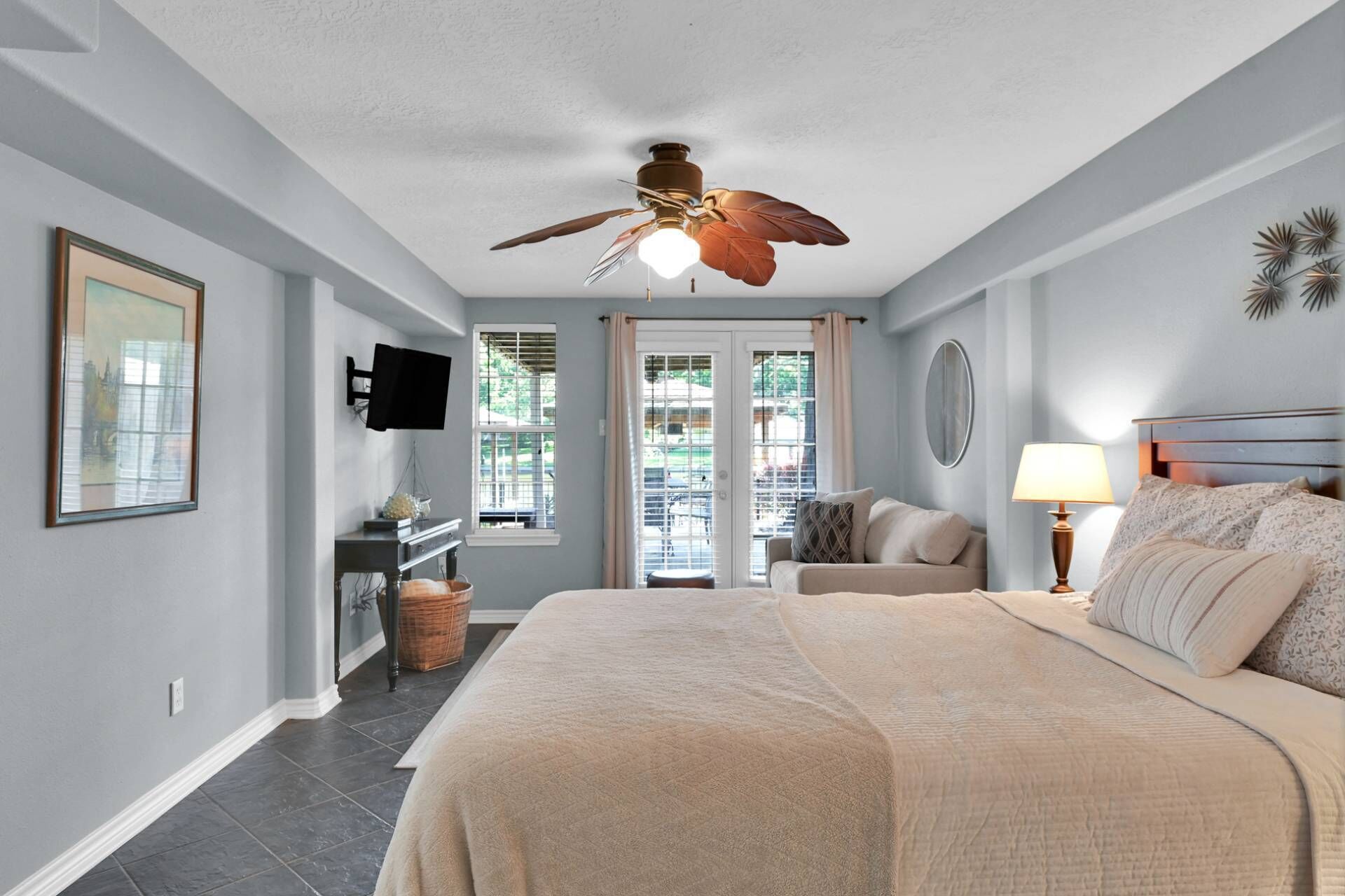 A bedroom with a bed, a ceiling fan, a desk with a chair, a TV mounted on the wall, and a window with blinds.