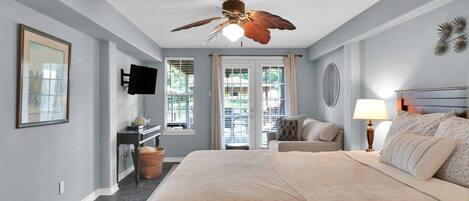 A bedroom with a bed, a ceiling fan, a desk with a chair, a TV mounted on the wall, and a window with blinds.