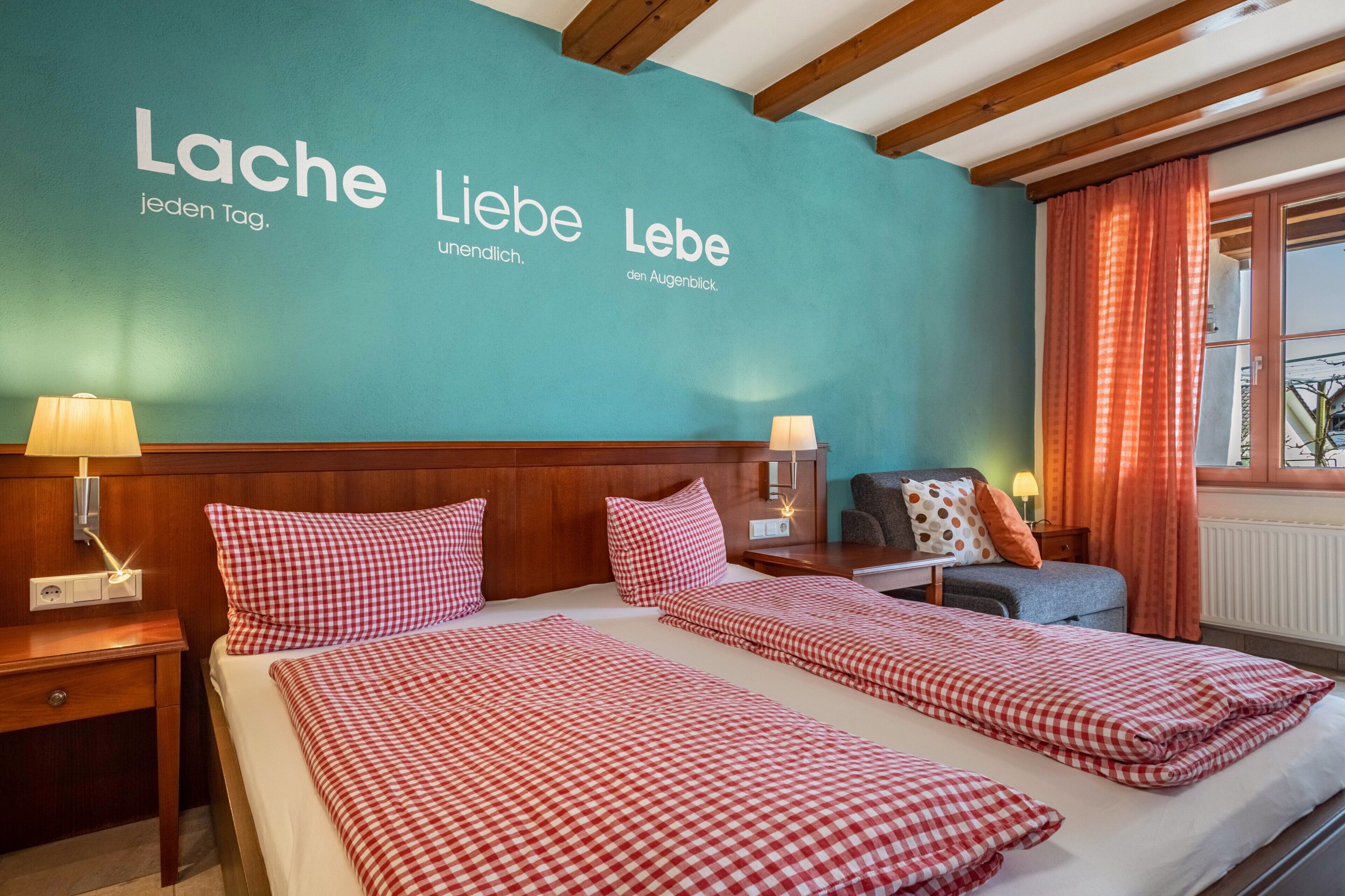 Een hotelkamer met twee bedden, een wand in turquoise, houten balken, een raam met gordijnen en een nachtkastje met een lamp.
