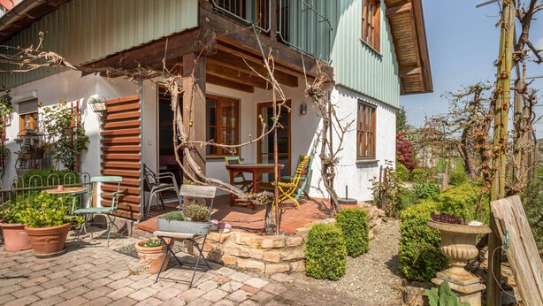A two-story house with a balcony, outdoor seating area with chairs and a table, and a garden with potted plants. - Room 'Kräutergärtle' with Private Terrace, Shared Garden and Wi-Fi (Immenstaad am Bodensee)