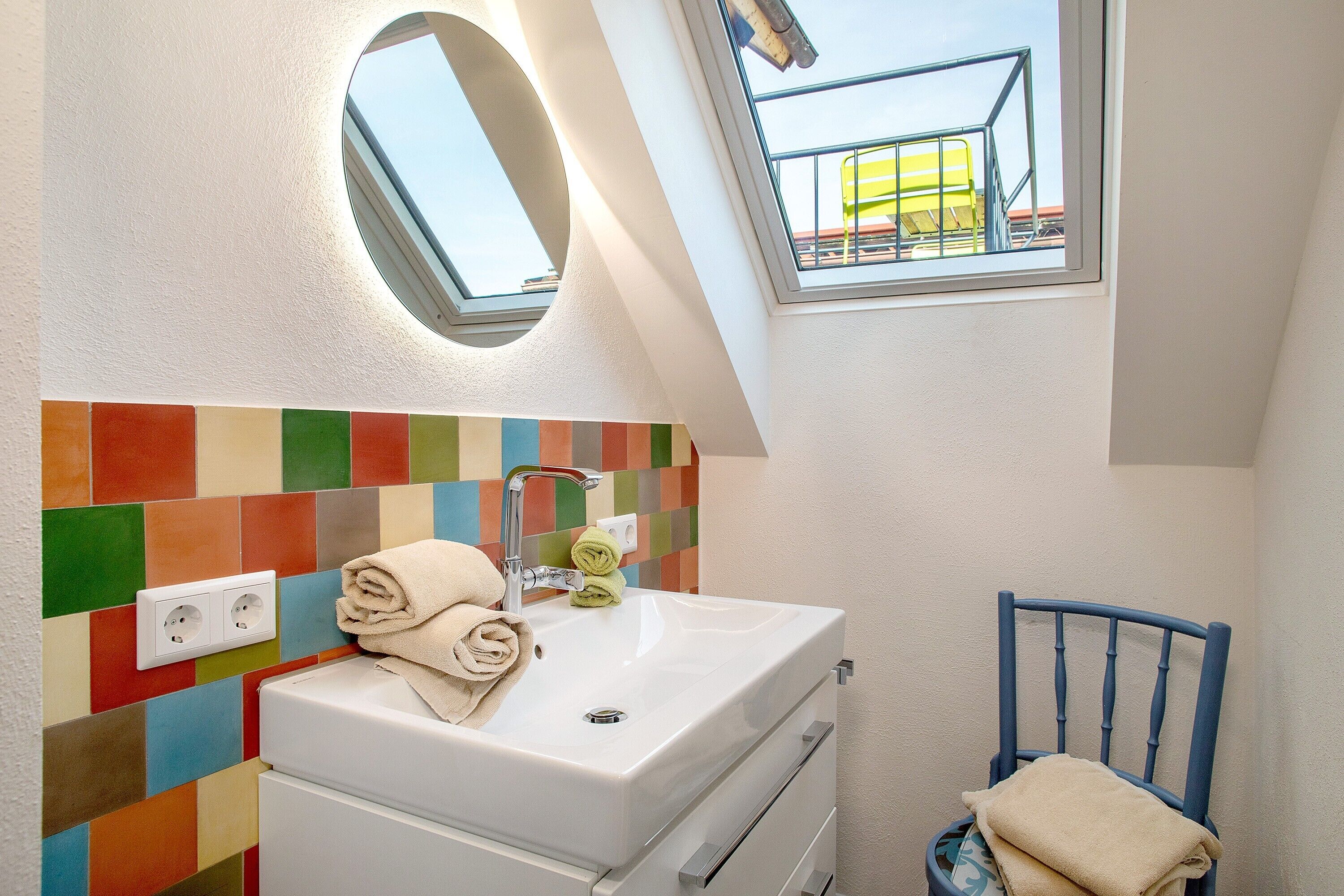 Un baño con un lavamanos blanco, un espejo, una pared de azulejos coloridos, una silla azul y un tragaluz.