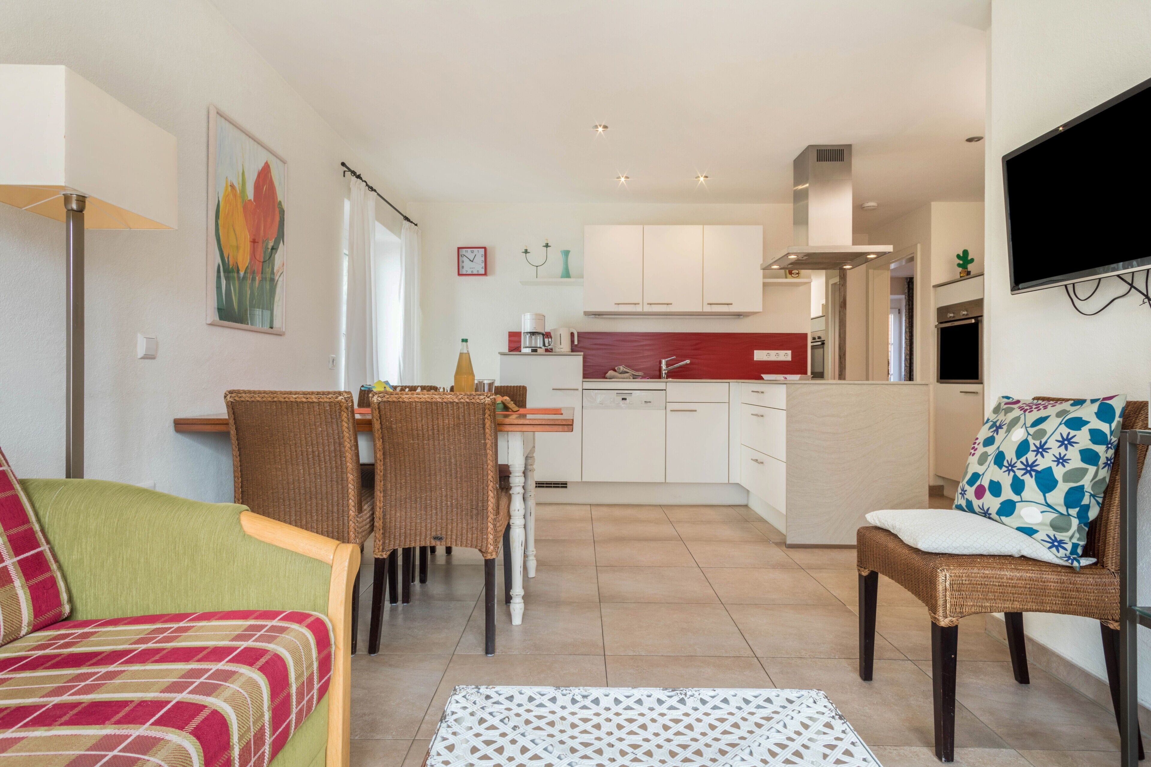 Een moderne keuken met witte kasten, een eethoek met rieten stoelen en een flatscreen tv aan de wand.