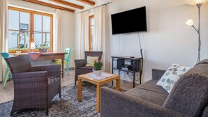 Una sala de estar con un sofá, dos sillas, una mesa con una planta, un televisor en la pared y una zona de comedor con una mesa y sillas.