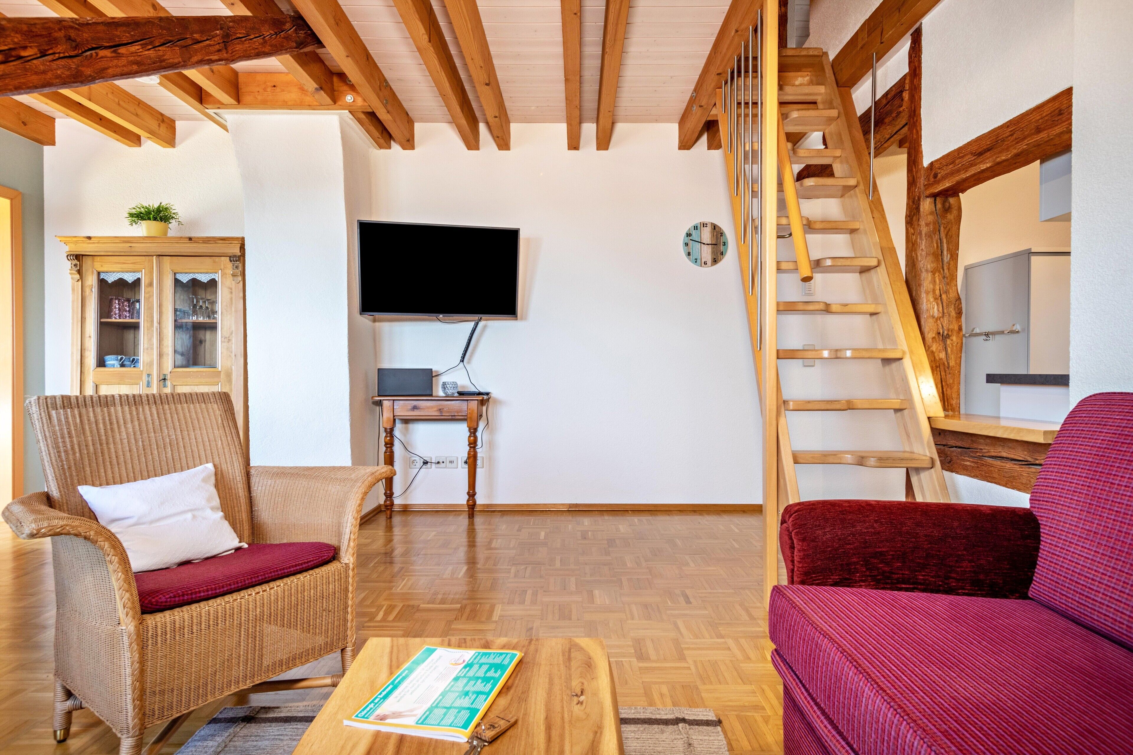 A living room with a wooden ceiling, a television, a wooden ladder, a wicker chair, a red sofa, and a wooden table.