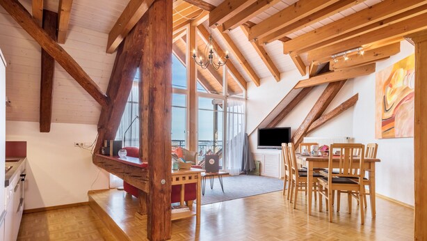 Spazioso loft con soffitti alti, travi a vista in legno, zona pranzo con sedie di legno e un'ampia finestra che offre luce naturale.