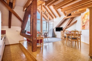 A spacious loft with high ceilings, exposed wooden beams, a dining area with wooden chairs, and a large window providing natural light.