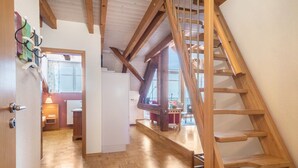 Una scala in legno conduce a un piano superiore, con un pavimento a motivi a spina di pesce, e si apre su una sala da pranzo con tavolo e sedie.