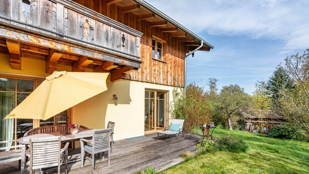 Een houten huis met een balkon, een terras met een tafel en stoelen, en een gazon.