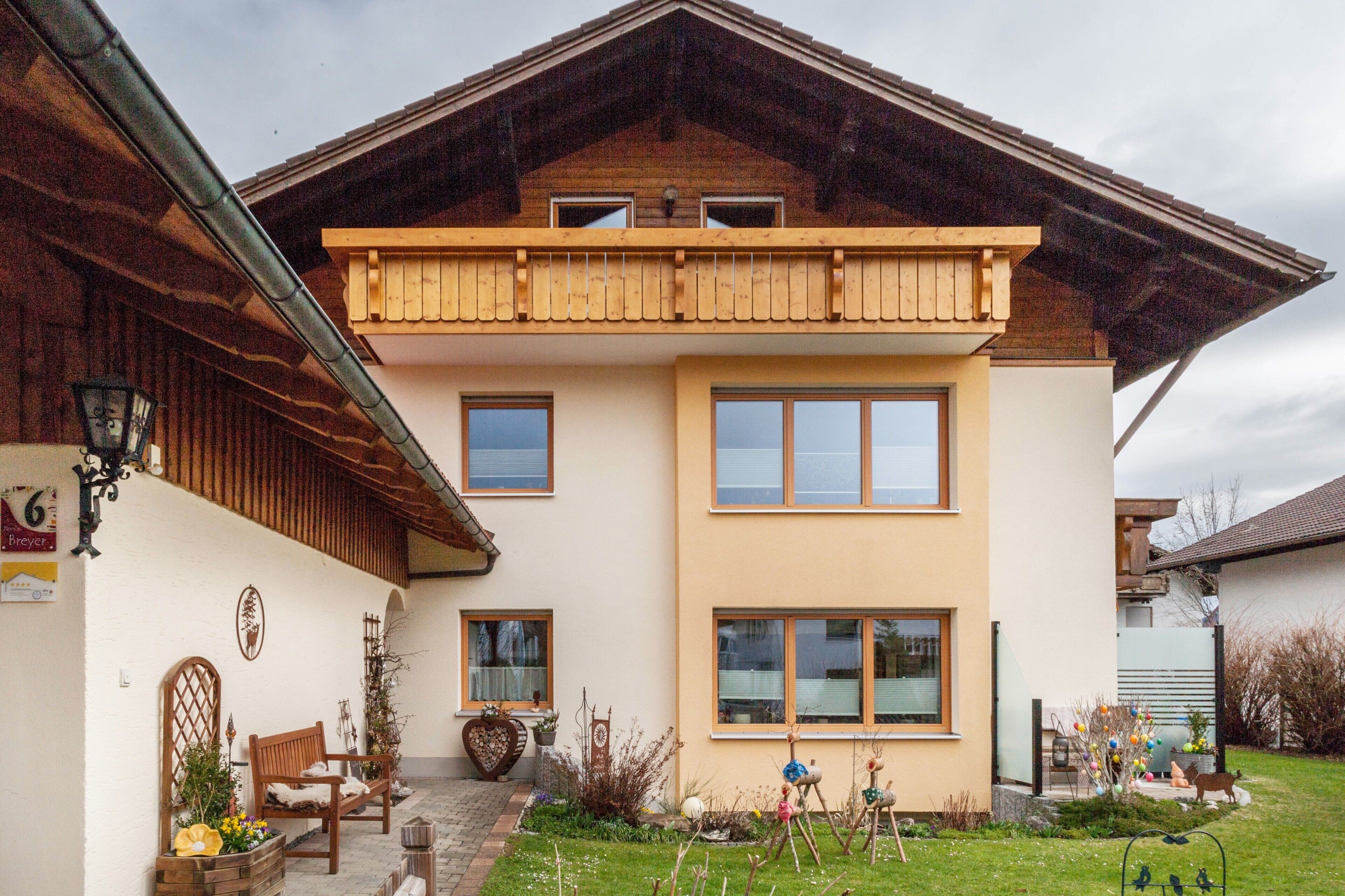A two-story house with a balcony, a garden with plants and a bench, and a wooden fence.