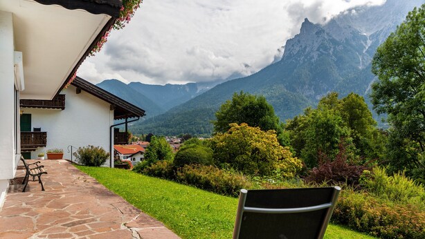 A stone-paved patio with a bench, a white building with a red roof, and a mountainous landscape in the background.