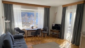 A modern living room with a sofa, dining table, chairs, a TV, and large windows with curtains.