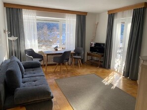 A modern living room with a sofa, dining table, chairs, a TV, and large windows with curtains.