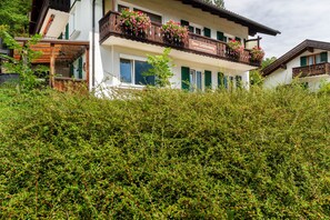 A two-story building with a balcony adorned with flowers, a sign indicating it is a guesthouse, and a lush green hedge in the foreground.