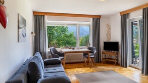 A living room with a sofa, a table, a TV on a stand, and a view of greenery outside.