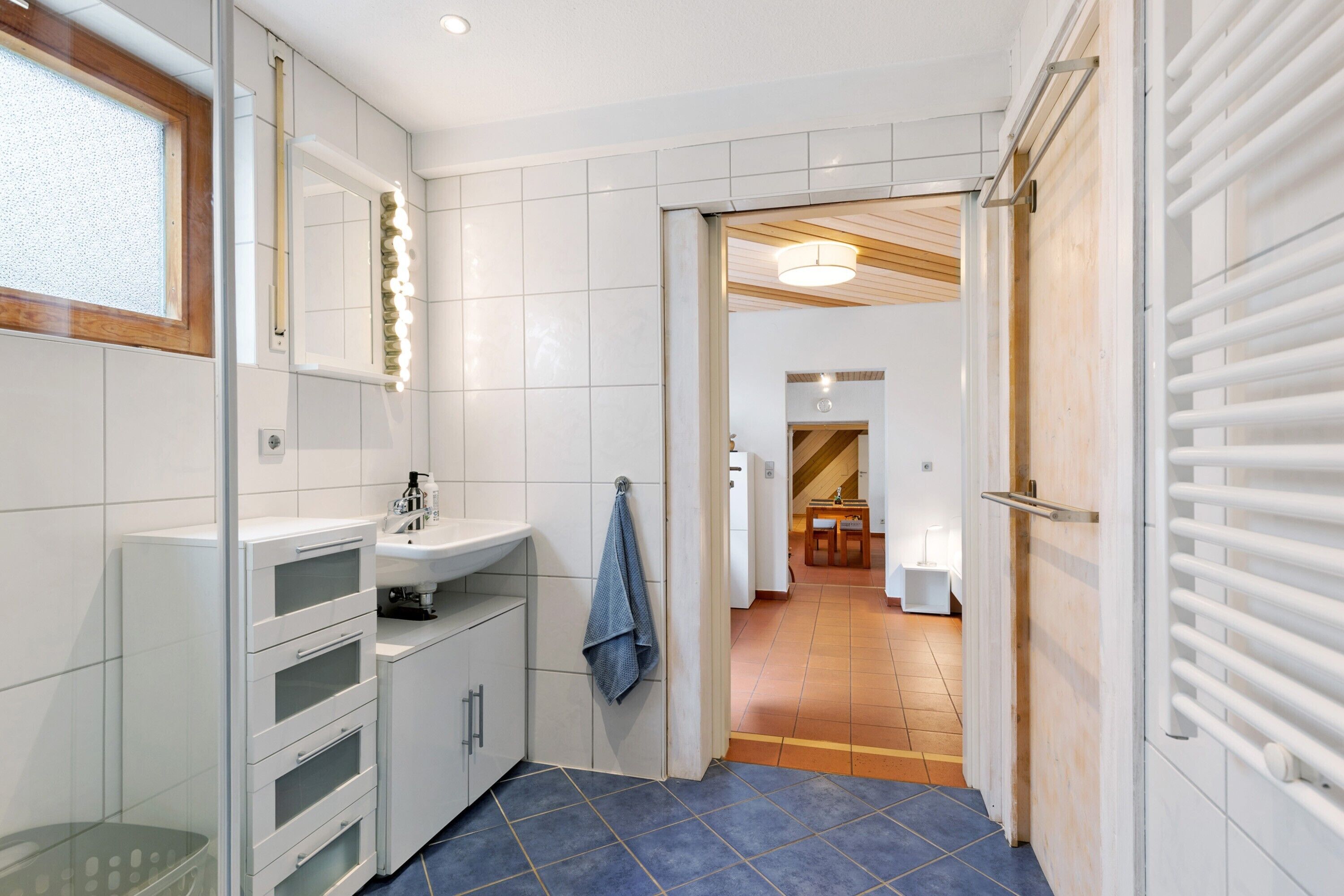 A narrow hallway with a bathroom on the left, featuring a sink, mirror, and towel rack. A wooden door leads to a dining area with a table and chairs.