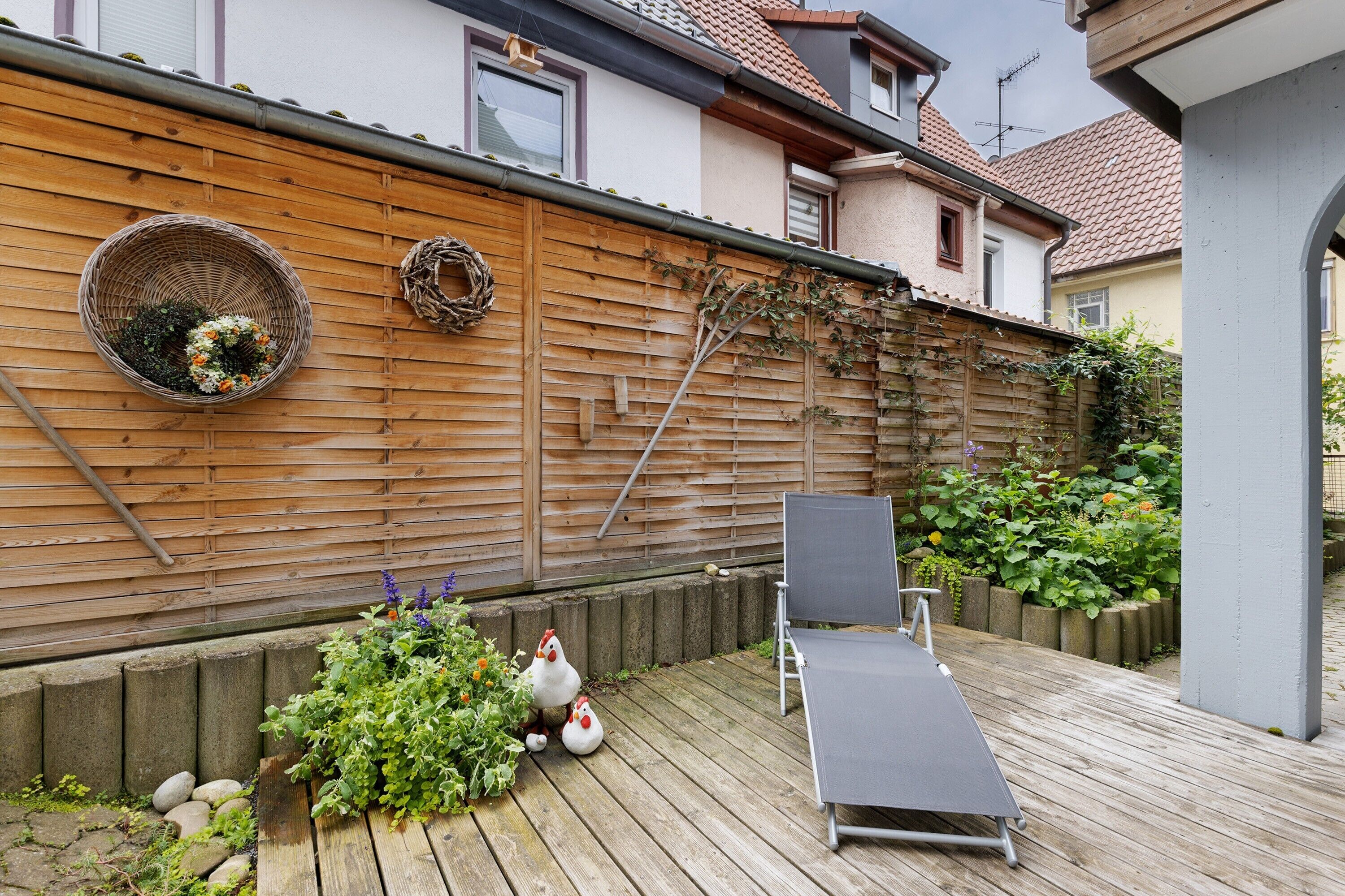 A wooden deck with a lounge chair, a wicker basket with flowers, and a garden wall.