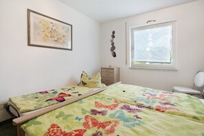 Quarto com duas camas, janela com persianas, mesa de cabeceira, uma cadeira e uma obra de arte na parede.