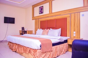 A hotel room with a large bed, a wooden headboard, a flat-screen TV mounted on the wall, a small desk, and a chair. - Owee Hotel Exclusive Apartments & Suites (Lagos)