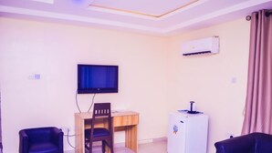 A small living room with a TV, a desk, a chair, a refrigerator, and a couch. - Owee Hotel Exclusive Apartments & Suites (Lagos)