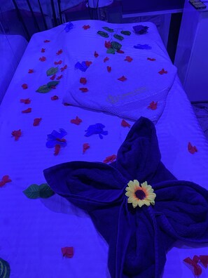 A room with a bed covered in a white sheet with a floral pattern, a dark blue towel with a sunflower, and scattered red and blue paper flowers. - Owee Hotel Exclusive Apartments & Suites (Lagos)