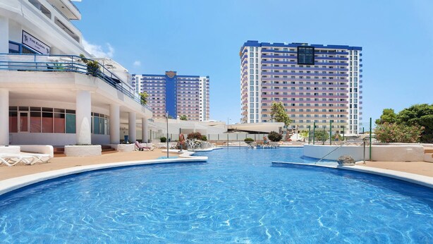 Una piscina con sillones de descanso y un edificio con balcones al fondo.