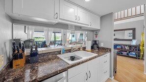 Eine moderne Küche mit weißen Schränken, einer Granitabdeckung und einem Spülbecken. Durch ein mit Vorhängen drapiertes Fenster ist ein Essbereich zu sehen.