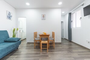 Een moderne woonkamer met een blauwe bank, een houten eettafel met stoelen, een keuken in de achtergrond en een groot raam met gordijn.