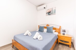 Een net opgemaakt bed met blauw beddengoed en twee opgerolde handdoeken, een nachtkastje met een lamp, en een wandairconditioner.