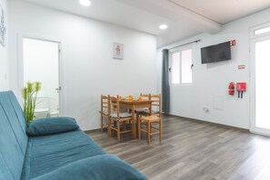 A room with a blue sofa, a wooden dining table with chairs, a TV mounted on the wall, and a fire extinguisher.