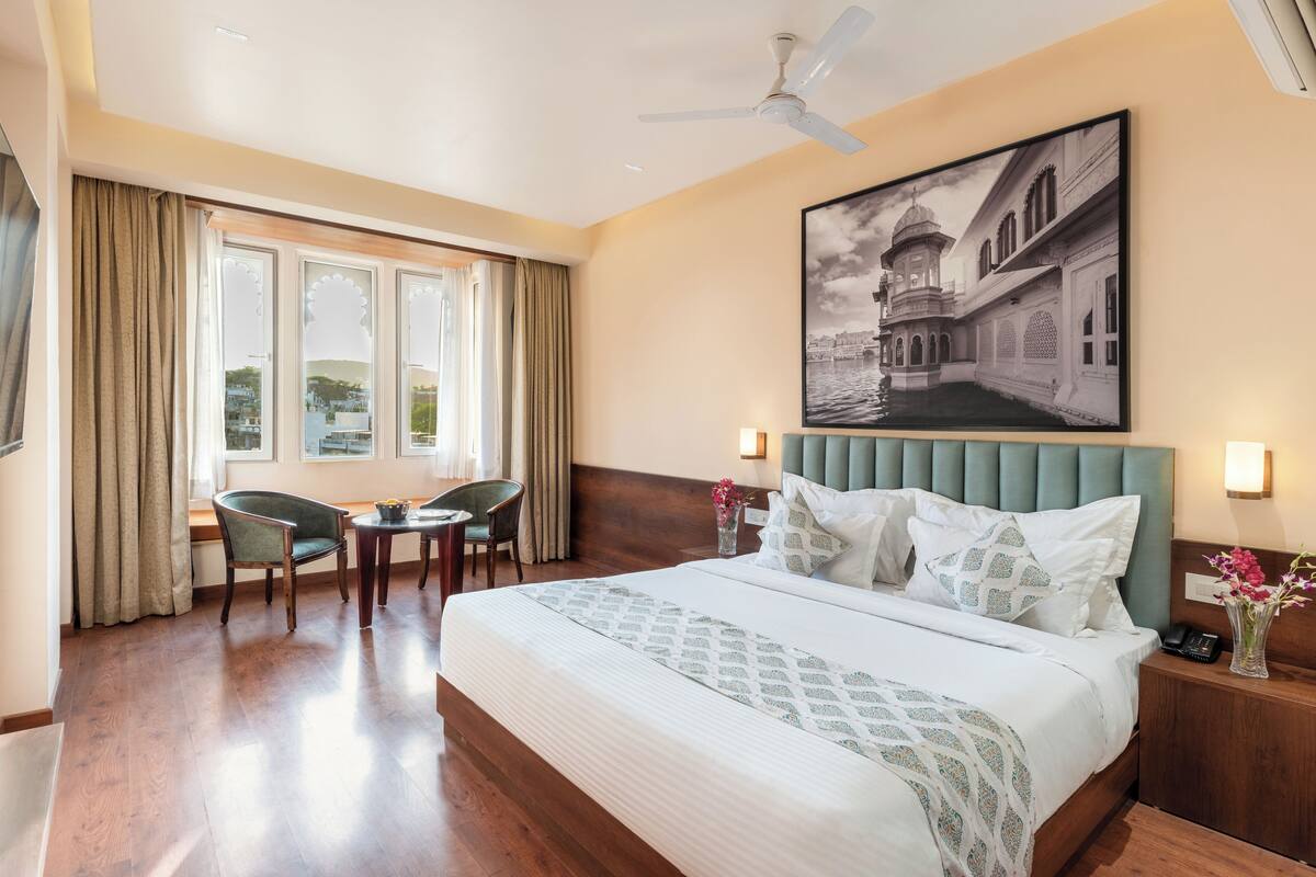 A hotel room with a large bed, a desk, two chairs, a ceiling fan, and a wall-mounted picture.