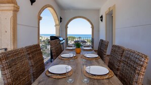 A dining area with a wooden table set for six, surrounded by wicker chairs, with a view of the sea through arches. - Castillo de Troya - Denia, Costa Blanca (Denia)