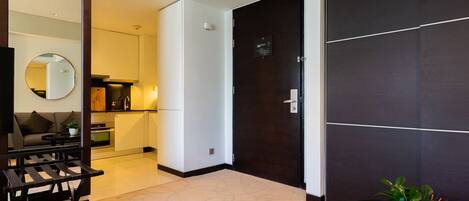 A modern hotel room with a dark wood door, a round mirror, a sofa, a dining table, and a kitchen area visible through an open door.