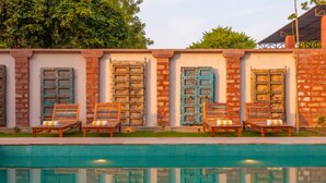 Zona junto a la piscina con piscina empavedada, sillas de madera con cojines y pared de ladrillo con contraventanas decorativas.
