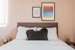 A neatly made bed with white and dark pillows, a framed inspirational quote, and a colorful abstract painting on the wall.