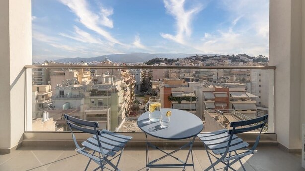 A balcony with a table and chairs, overlooking a cityscape with buildings and mountains in the distance. - Exceptional Living Loft 801 (Athina)