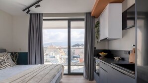 A modern bedroom with a large bed, a view of the city through a sliding glass door, and a kitchenette with a refrigerator and sink. - Exceptional Living Loft 801 (Athina)