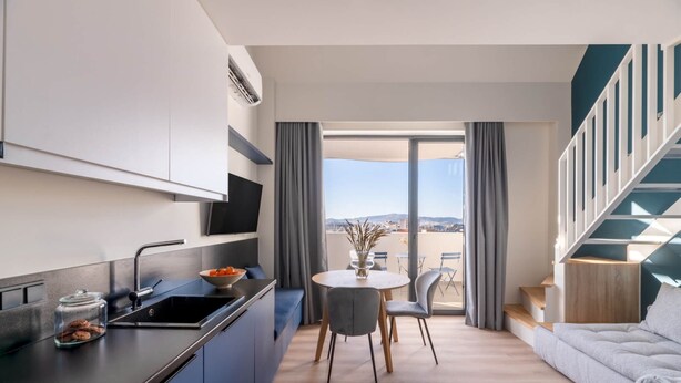 A modern kitchen with a built-in stove, a dining area with a round table and chairs, and a view of the outdoors through large windows. - Exceptional Living Loft 704 (Athina)