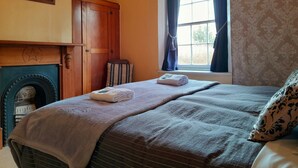 A bedroom with a bed, a fireplace, a window with curtains, and a patterned wall. - Tregonning Cottage Waipiata (Waipiata)