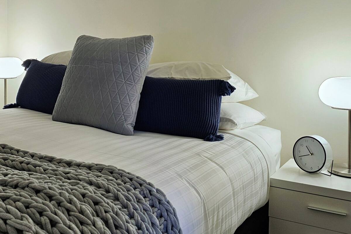 Un lit bien fait, avec une couette grise et un plaid épais en tricot, deux lampes de chevet, une horloge ronde fixée au mur et une petite commode.