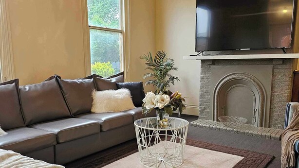 A living room with a sofa, a coffee table with a vase, a fireplace, and a TV mounted above it. - Bendigo Holiday Accommodation (Bendigo)
