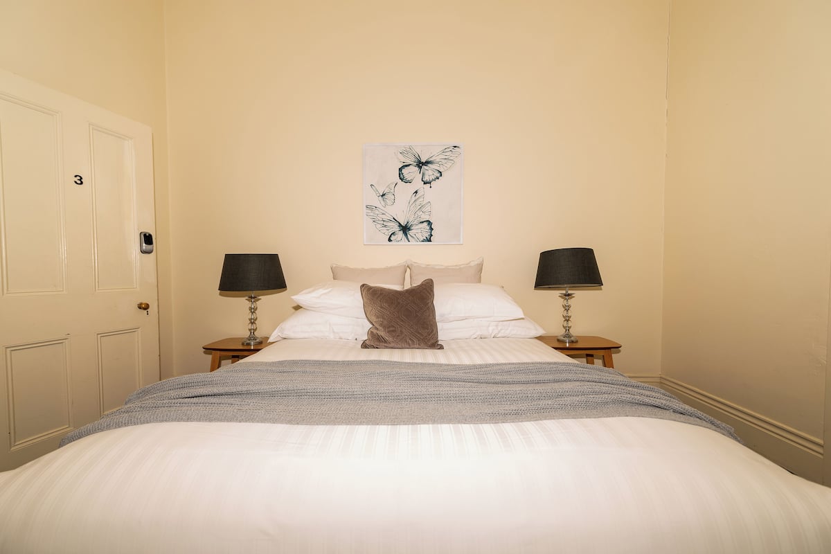 Un lit bien fait, recouvert d’une couverture grise, avec deux lampes de chevet et une œuvre d’art représentant des papillons, accrochée au mur.