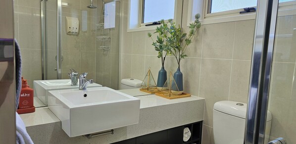 Un baño con dos lavamanos, una cabina de ducha de vidrio y un inodoro. Hay jarrones decorativos en el mostrador.