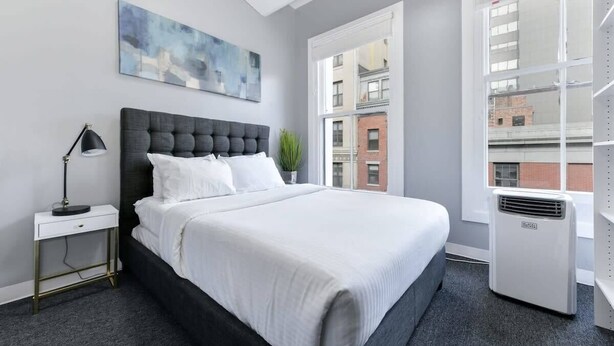 A modern bedroom with a large bed, a bedside table with a lamp, a potted plant, and a large window with a city view. - Downtown Crossing, Affordable Stay, Book Now! (Boston)