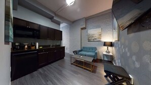 A modern kitchen with dark cabinets, a microwave, and a coffee machine. There is a blue sofa and a small table with a lamp. The room has a gray brick wall and a painting above the sofa. - Downtown Crossing, Affordable Stay, Book Now! (Boston)