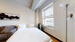 Une chambre à coucher comprenant un lit, un canapé, une fenêtre avec des stores, une lampe sur pied et un téléviseur fixé au mur.