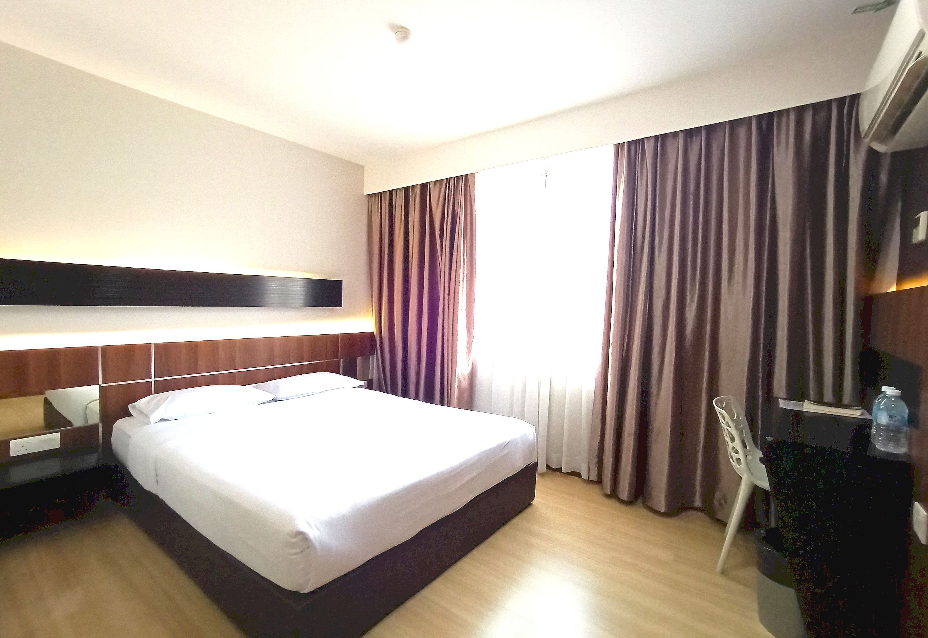 A hotel room with a large bed, a wooden headboard, a desk with a chair, and a window with curtains.