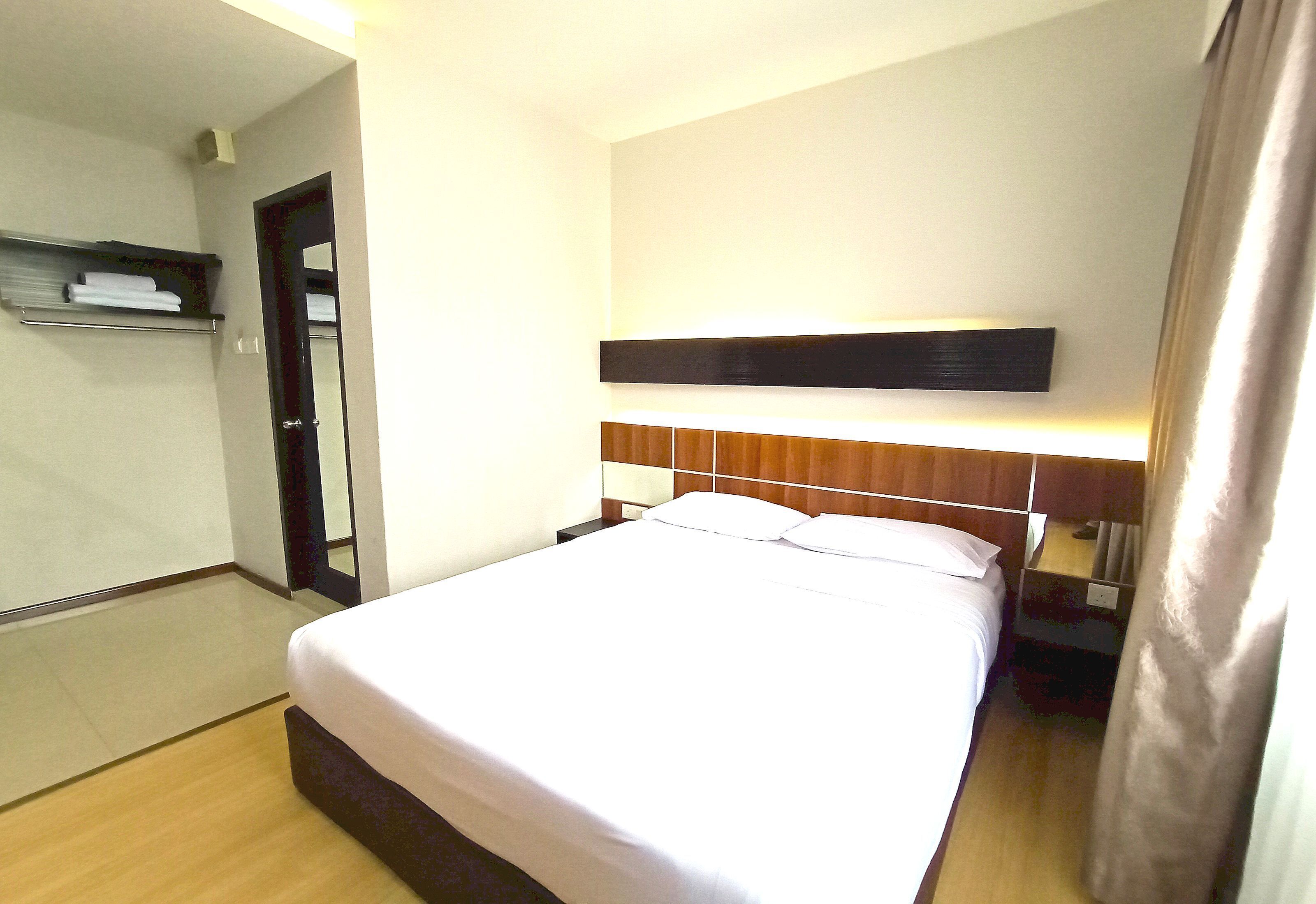 Una habitación de hotel con cama, mesita de noche, repisa con toallas y una puerta.