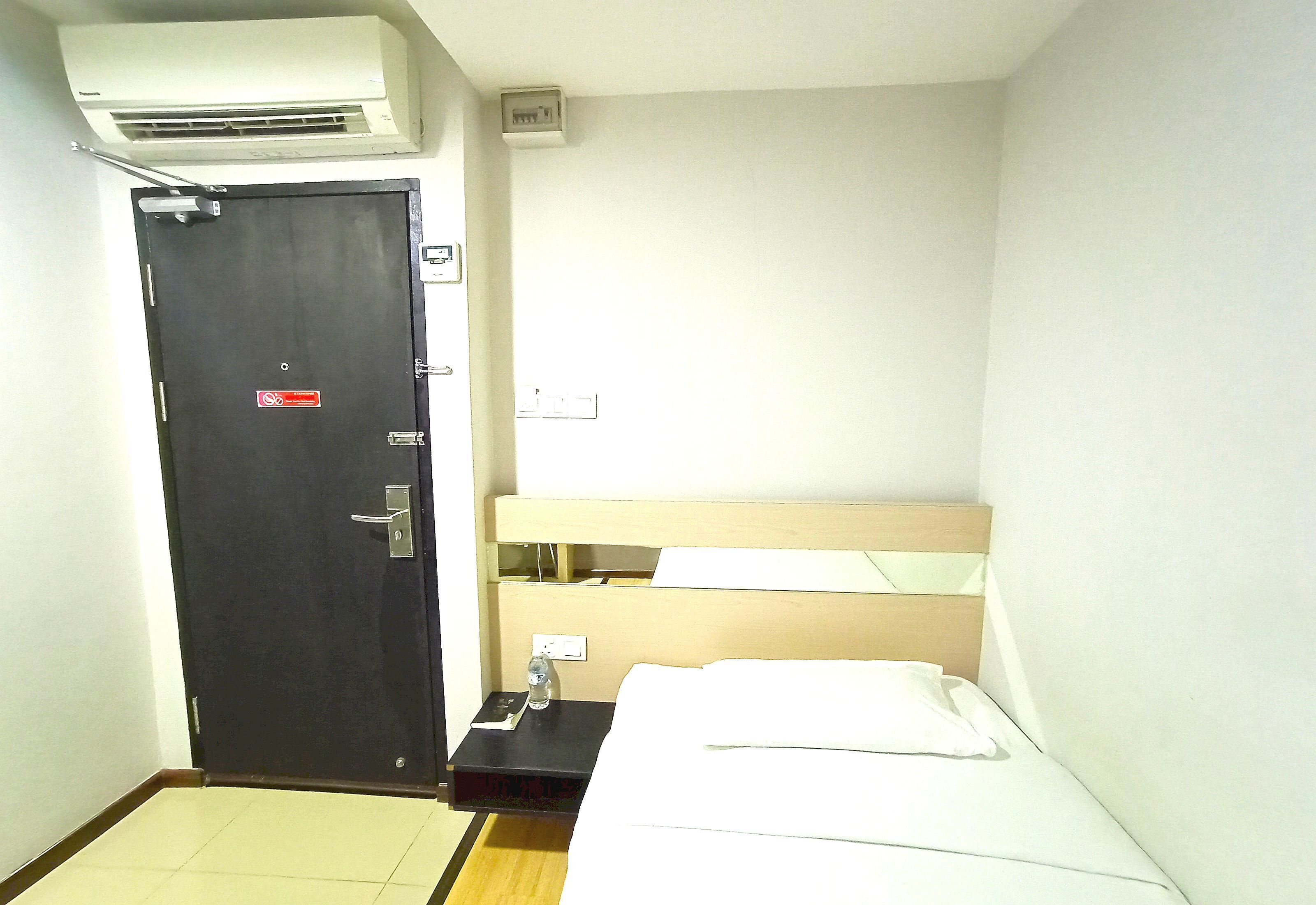 Una habitación de hotel pequeña con una cama individual, una mesita de noche, una puerta y un equipo de aire acondicionado.