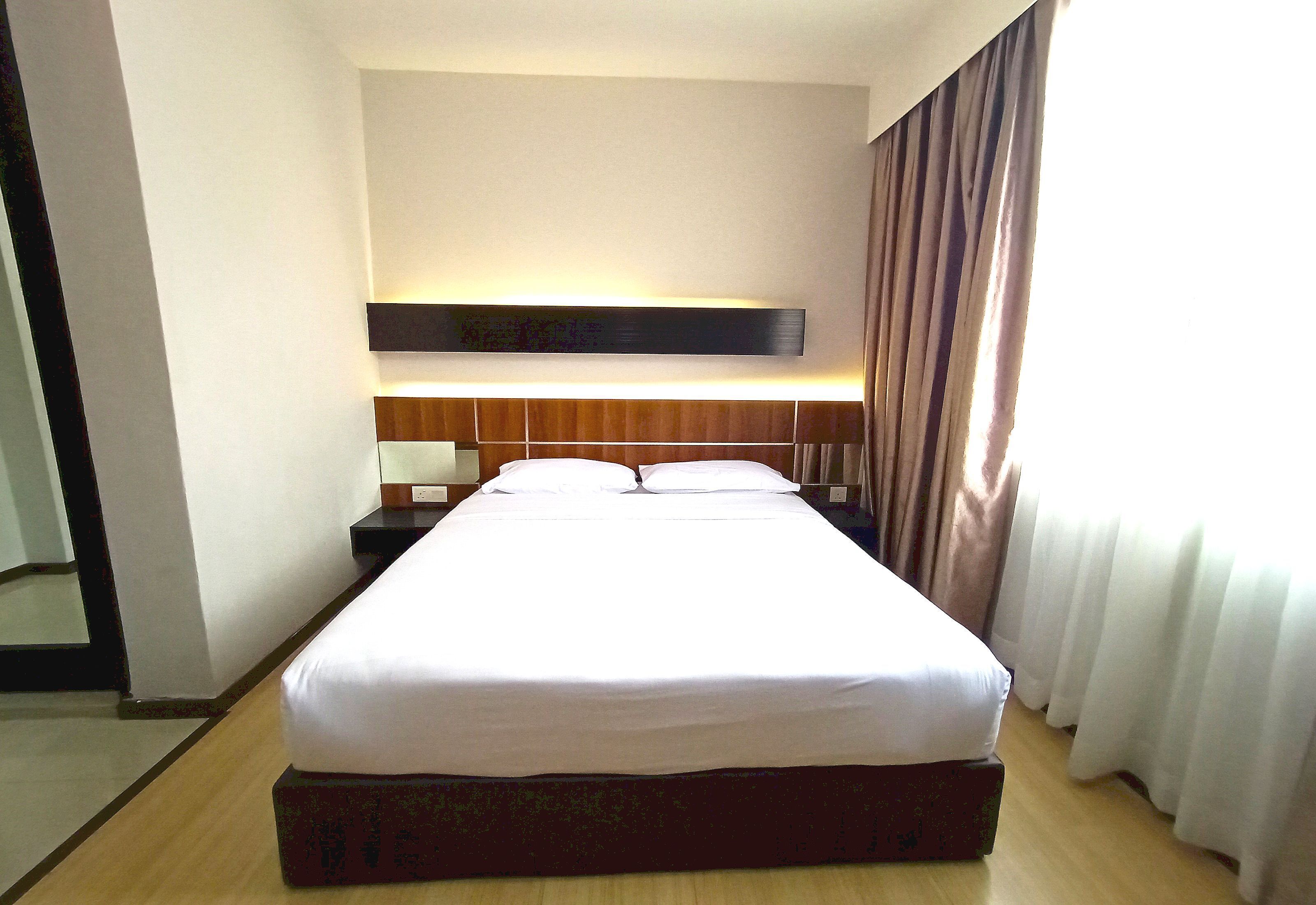 A hotel room with a large bed, a wooden headboard, a bedside table with a lamp, and a window with curtains.